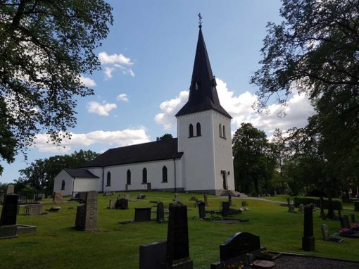 Baksidan av Hjälstads kyrka med lutande kyrkogård och flera gravar, omsluten av frodig grönska.