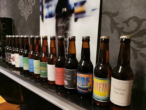 En rad olika ölsorter från Åsalid Bryggeri Falköping uppställda på en svart hylla, med etiketter som representerar bryggeriets unika smaker.