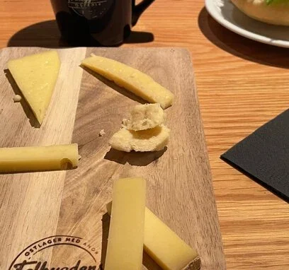 Falbygdens Turism – Smakupplevelser på Falbygdens Osteria En ostbricka med olika ostar på ett träfat märkt med Falbygdens Osteria, tillsammans med en kopp kaffe och en fralla i bakgrunden.
