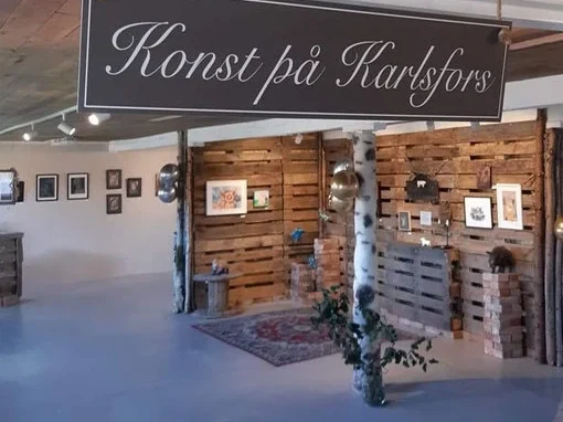 Interiör av Konst på Karlsfors med skylt över ingången och rustika detaljer i trä, omgiven av konstverk på väggarna.