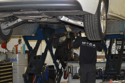 MECA Ingemarssons Bilservice AB - Verkstad Bild av en bil på lyft hos MECA Ingemarssons Bilservice AB, där en mekaniker arbetar under bilen i verkstaden.