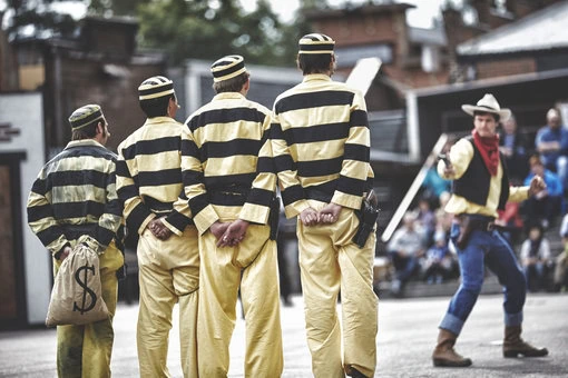 High Chaparral – sheriffer och skurkar Fyra skurkar i randiga fångdräkter står på rad medan en cowboy riktar sin pistol mot dem i en uppvisning på High Chaparral.