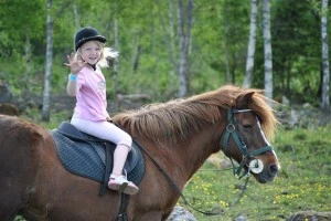 Hällstad Islandshästar – barn och unga Ett glatt barn i hjälm rider en brun islandshäst i en grön skogsmiljö.
