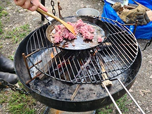Hällstad Islandshästar – mat i skogen En närbild på ett utomhuskök där mat tillagas över öppen eld, med grillverktyg och råvaror runt elden.