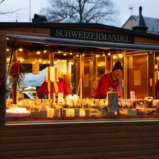 En julbod med skylten "Schweizermandel" på en traditionell julmarknad. Två personer i röda kläder säljer rostade nötter och julgodis i en träbod med varm belysning och dekorationer.
