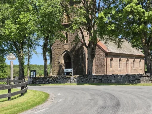 Vy mot kyrkporten i röd granit vid infarten till Vistorps kyrka, omgiven av träd och stenmurar.