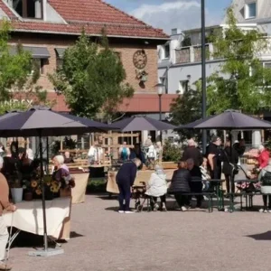 Besökare promenerar och sitter vid bord under parasoll på Malta Johanna Marknad i Falköping, med försäljningsstånd fyllda med blommor och lokala produkter.