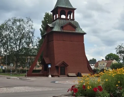 Röd klockstapel i trä vid Mora kyrka med kopparklätt torn, omgiven av blommor och grönska.