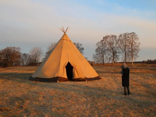 Tipitält vid Hornborgasjön under morgongryningen Ett tipitält står uppställt i det mjuka morgonljuset vid Hornborgasjön medan en besökare fotograferar den vackra soluppgången.