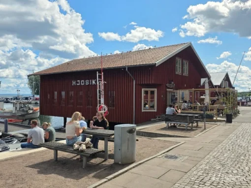 Hamnboden i Hjo med röd byggnad vid hamnen, uteservering, sittplatser, båtar och Vättern i bakgrunden.